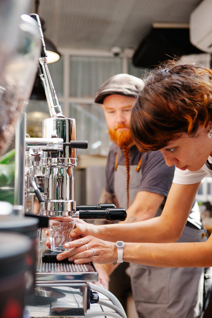 Focused baristas with glass using coffee machine to brew coffee in professional coffeehouse with special equipment during work process on blurred background